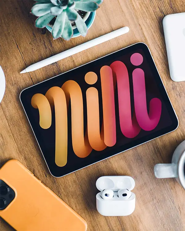 Ein Tablet, auf dem bunte "Mini"-Texte angezeigt werden, zusammen mit einem Stylus, einem Smartphone, Kopfhörern und einer Pflanze auf einer Holzoberfläche. Darauf sind individuelle Icons und Illustrationen zu sehen.